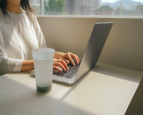 A person pouring water into a glass next to a laptop on a desk
